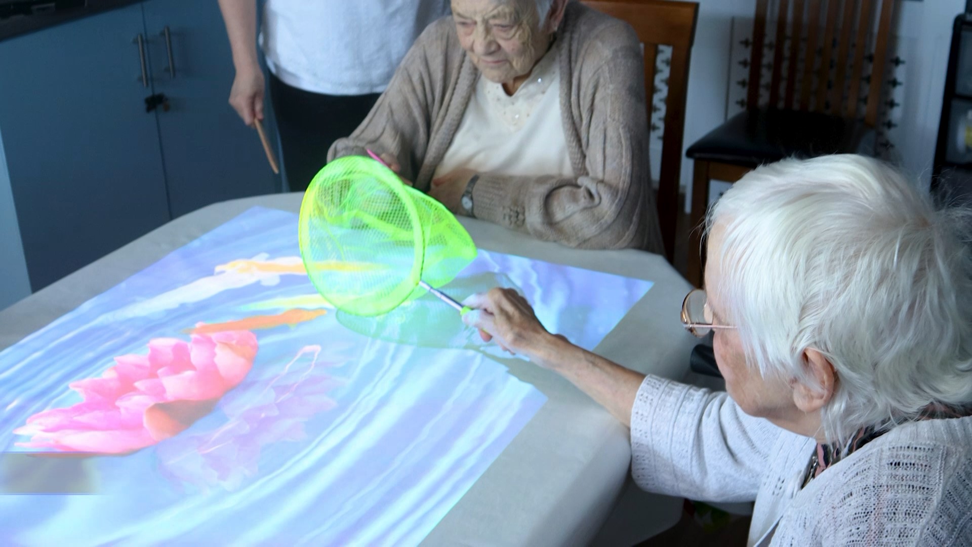 Two elderly ladies are seated at a table, interacting with the interactive fish pond being displayed from the interactive projector, the women are happily trying to catch the fish with a fishing net.