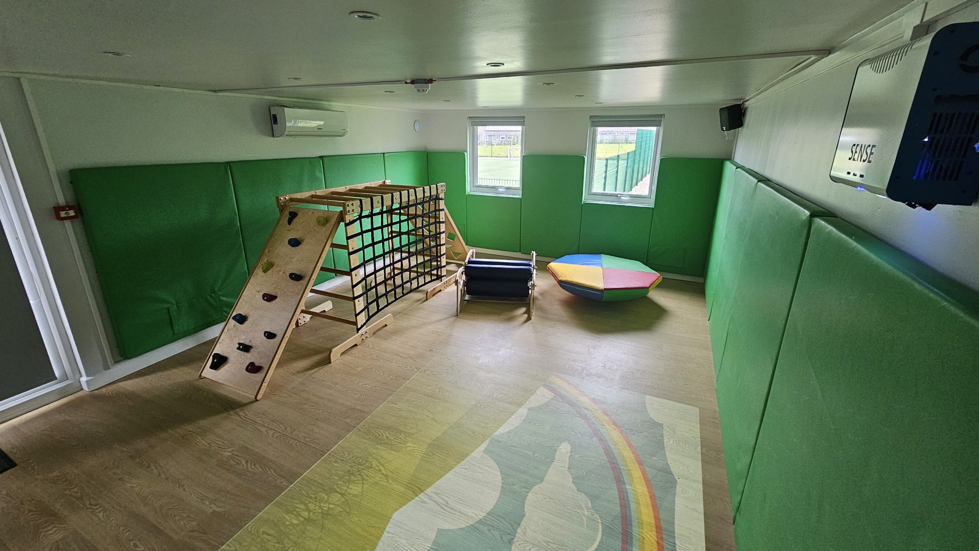 The image shows an example of a Sensory integration room within a school environment. The Sensory Integration room features a climbing system to promote strength, balance and coordination while providing a fun and engaging sensory activity. The Sensory Integration room has green soft padding to the walls for safety. The Sensory room features a soft padded rocker roller, a sensory body roller and a SENse Air wall mounted interactive sensory floor projection.