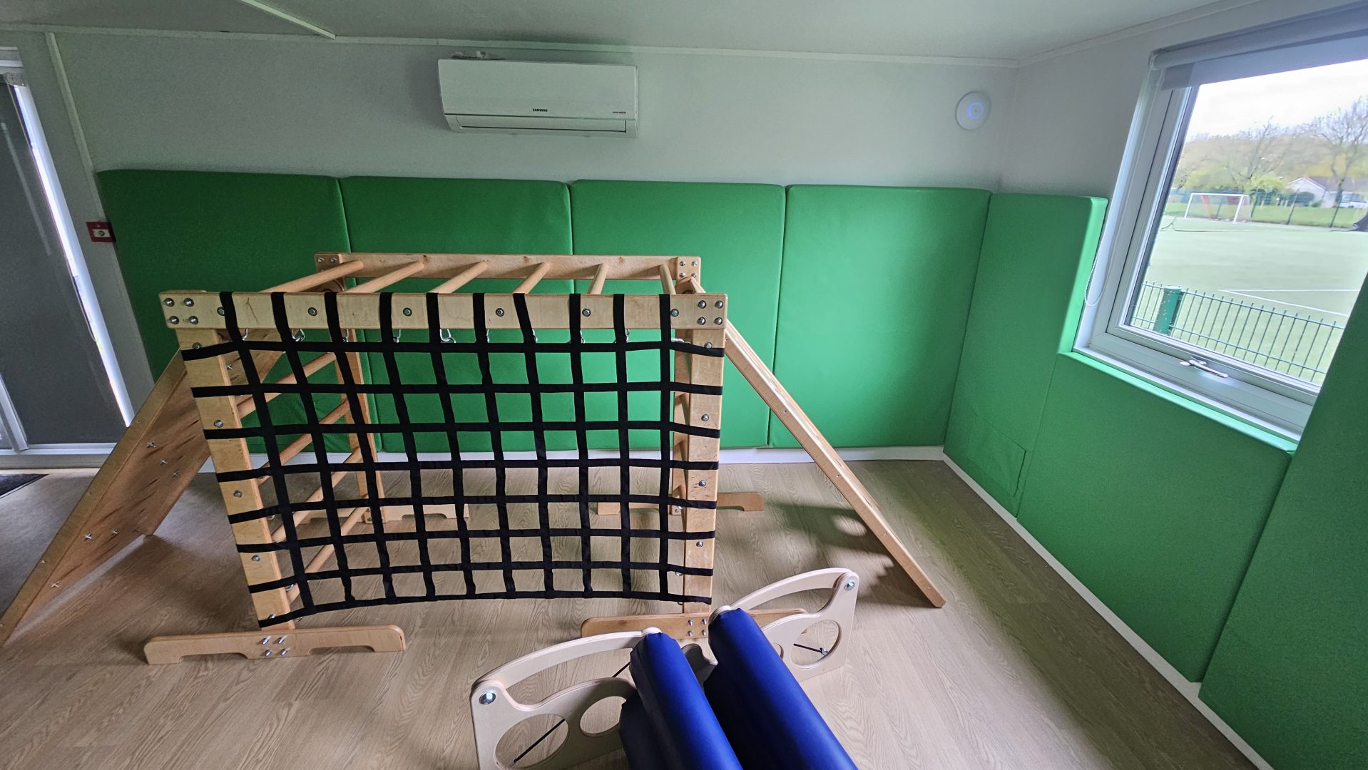 An image of the sensory climbing frame installed into a sensory integration room, the room also has a sensory therapeutic body roller and green soft padded walls. The room has a window providing natural light into the room
