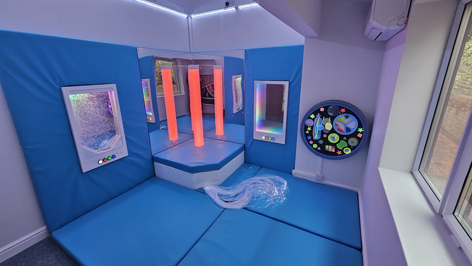 A well-equipped sensory room featuring calming chroma tubes, fibre optic lights, and interactive panels. The room has blue padded walls and flooring for safety, with a variety of sensory tools designed to help users with sensory processing disorders. Large windows allow natural light to complement the soothing environment. The Snow-Scape interactive panel allows users to immerse themselves in the beauty and joy of snowfall all year round. The Infinity-Glow Interactive panel provides an enchanting beauty of infinite reflections.