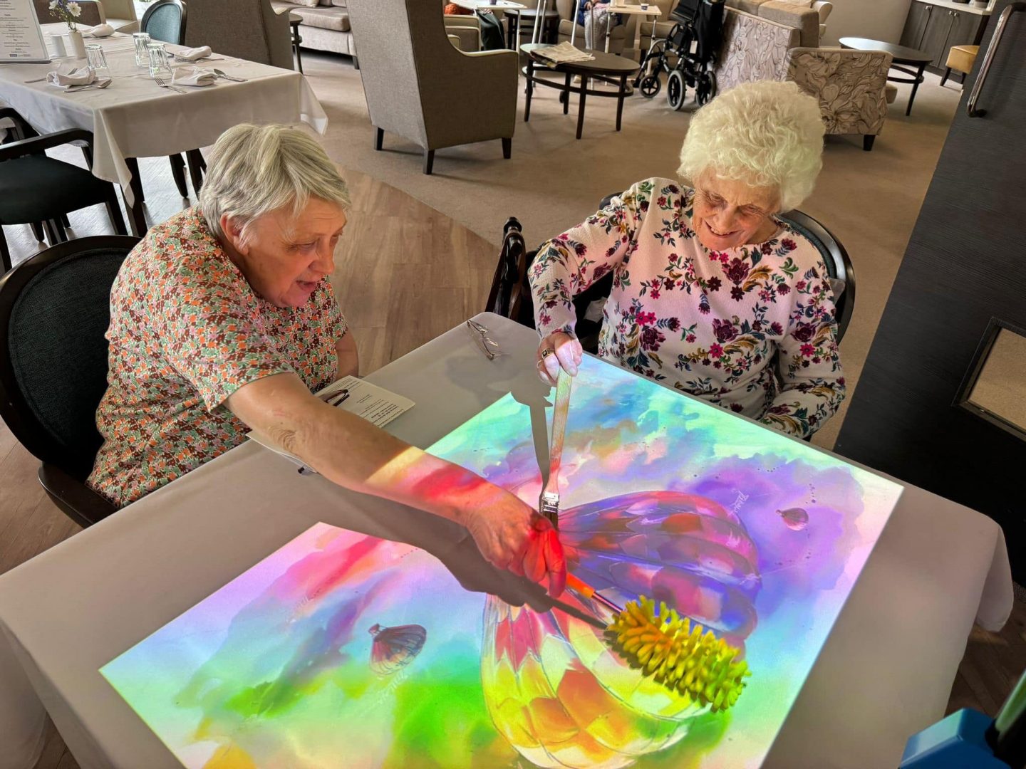 Two elderly ladies are enjoying a colouring in activity on the interactive projection table, the women are using dusters and paintbrushes to turn the image of the hot air balloon from black and white to a vibrant range of colours.