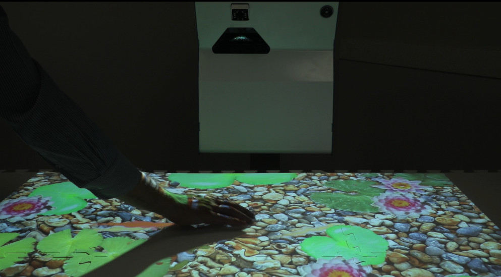 The SENse Flex interactive projector being used as an interactive table to display an interactive fish pond onto the table, this image is being interacted with a hand and the water ripples with movement.