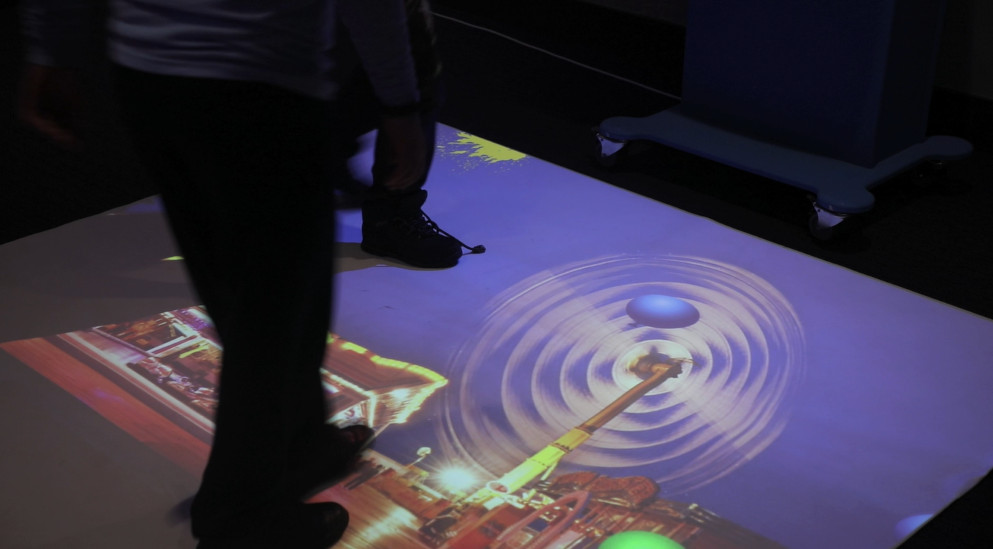 Bright interactive floor projector, with two people splatting the flying balloons, with a fairground background.