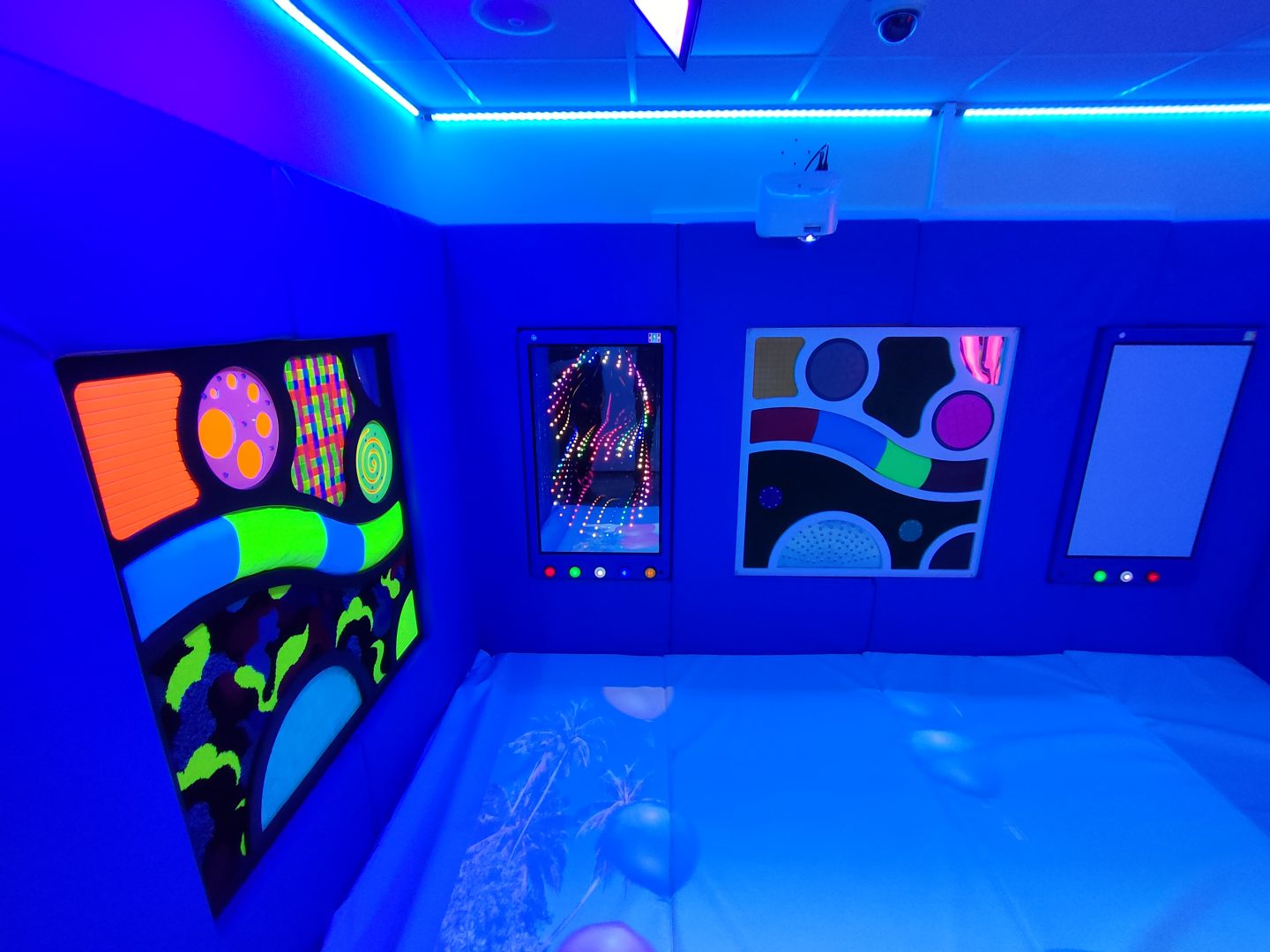 The image shows a sensory room illuminated with blue lighting, featuring interactive and colourful panels on the walls. The panels display various patterns and shapes, including spirals, abstract designs, and geometric figures that are vividly highlighted by the black light. There are also buttons with different colours at the bottom of each panel, likely used to interact with the panels. The floor is covered with a soft padded floor, creating a safe and engaging environment. The floor of the sensory room features an interactive projector.
