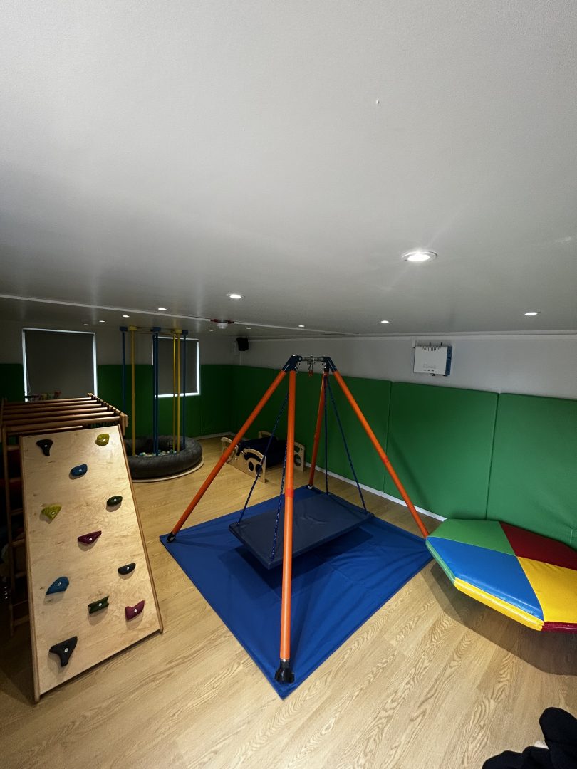 A well equipped sensory integration room, the users of this room have access to a large platform swing ideal for occupational therapy sessions, a climbing frame for training the sense of spatial orientation and balance and a soft padded rocker roller for the children to sit or climb onto. The room has green soft padded walls for safety.